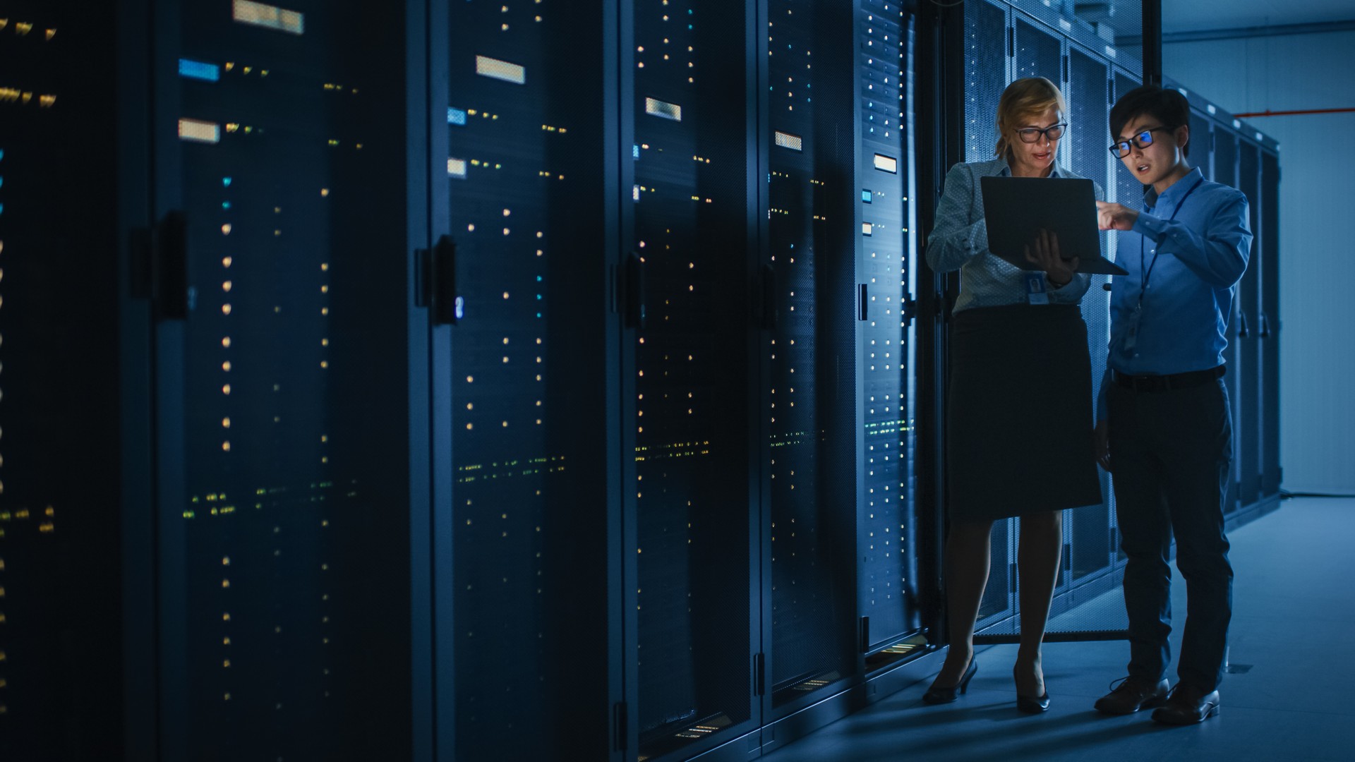 In the Modern Data Center: Engineer and IT Specialist Work with Server Racks, Talking, Doing System Maintenance Check with Laptop Computer. Concept of Diagnostics of the Database, Cybersecurity Check